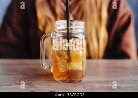 Ein Glas Eistee. Transparenter Behälter mit Strohhalm. Kaltes erfrischendes Getränk auf dem Tisch. Eiswürfel in bernsteinfarbener Flüssigkeit. Sommergetränk im Café. Entspannender Drink. Stockfoto