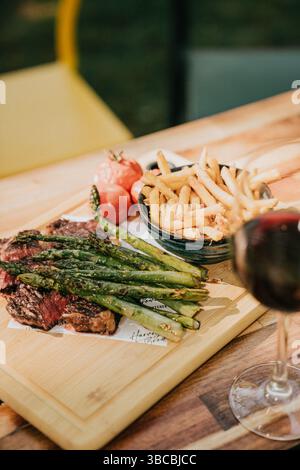 Ein Teller mit Steak und Spargel sitzt auf einem hölzernen Schneidebrett. Ein Glas Wein steht neben dem Teller Stockfoto