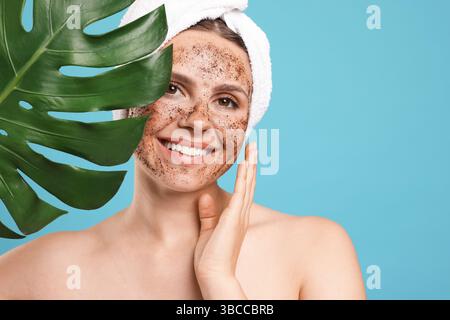 Lächelnde Frau mit Körperpeeling im Gesicht und Monstera-Blatt vor hellblauem Hintergrund Stockfoto