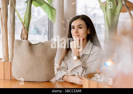 Stilvolle Frau, die sich in einem gemütlichen Café mit natürlichem Dekor entspannt, mit eleganter Uhr und Leinen-Outfit. Stockfoto