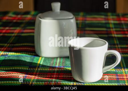 Eine helle und gemütliche Szene mit einer weißen Keramikkkaffeetasse und einer Zuckerschale auf einer farbenfrohen Tischdecke mit Schottenmuster. Das warme Sonnenlicht enhan Stockfoto