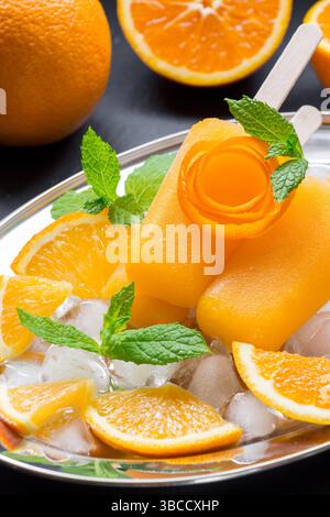 Orangensorbet-Eis dekoriert mit einer Blüte von Orangenschale auf Silberplatte mit Eiswürfeln, Orangenstücken, Minze in der Nähe von Orangenscheiben auf schwarzem Hintergrund. Orangensorbet-Eis-Eis-Eis. Vertikal. Stockfoto