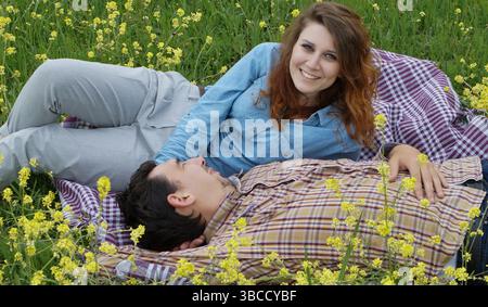 Ein junges Paar, das sich auf einer blühenden Wiese auf einer karierten Decke entspannt. Die Frau lächelt in die Kamera, während der Mann neben ihr ruht. Stockfoto