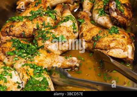 Gebackene Hähnchenschenkel in eigenem Fett, bestreut mit gehackter Petersilie und Knoblauch in einer Schüssel mit einer Zange Nahaufnahme. Stockfoto