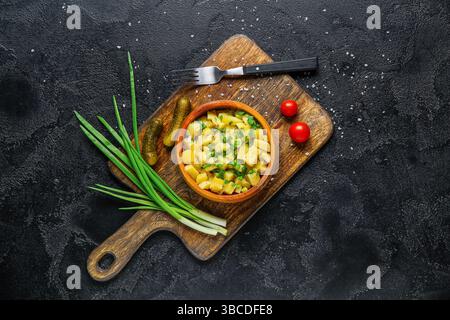Hölzerne Schüssel mit leckerem Kartoffelsalat mit grünen Zwiebeln, Tomaten und eingelegten Gurken auf schwarzem Hintergrund Stockfoto