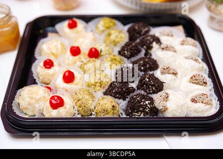 Elegantes Tablett mit verschiedenen Dessertbällchen mit Pistazien, Schokoladenstreuseln, Kokos, Kirschen und Sesam – ideal für Veranstaltungen oder Geschenke. Stockfoto
