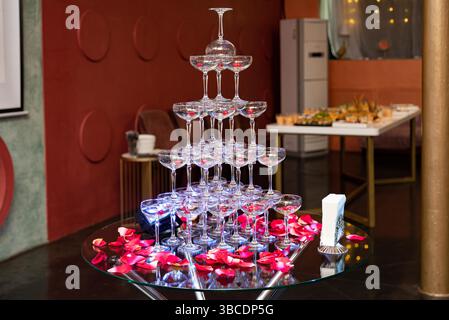 Eine Pyramide aus leeren Sektgläsern mit Kirschen und Rosenblättern, die für eine Feier oder eine formelle Veranstaltung eingerichtet sind. Stockfoto
