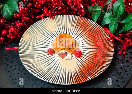 Festliches Dessert mit einem kleinen Kuchen, frischen Himbeeren und herzförmiger Schokolade, dekoriert mit rotem Dekor für eine Weihnachtsfeier. Stockfoto