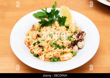 Ein farbenfroher Teller mit Reis mit Meeresfrüchten und grünen Erbsen, garniert mit Petersilie und Zitronenkeil, serviert auf einer weißen Keramikschale. Stockfoto