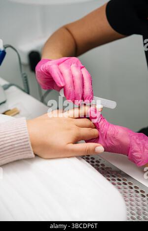 Nahaufnahme eines Nageltechnikers in rosa Handschuhen, der die Fingernägel eines Kunden während einer professionellen Maniküre in einem Salon feilt. Stockfoto