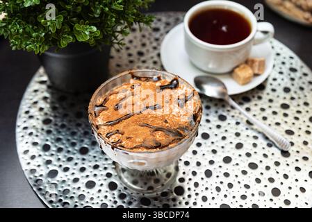 Klassisches italienisches Tiramisu, serviert in einer Glasschale, belegt mit Kakaopulver und Schokoladennebel, gepaart mit heißem schwarzem Kaffee. Stockfoto