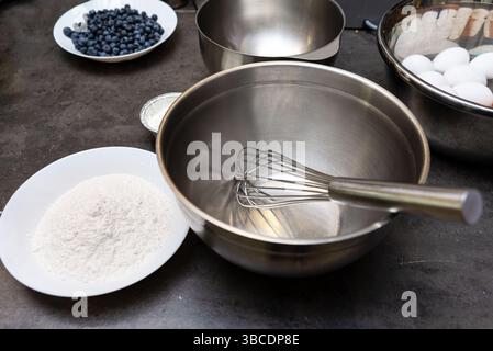 Rührschüssel aus Edelstahl mit Schneebesen, umgeben von Backzutaten, einschließlich Mehl, Eiern und frischen Heidelbeeren, bereit für die Rezeptzubereitung. Stockfoto