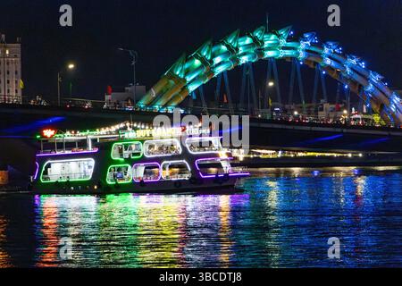 DA NANG, VIETNAM, 27. APRIL 2025, fährt Ein hell dekoriertes Flussboot am Abend auf dem Fluss mit der beleuchteten Drachenbrücke auf einem Hintergrund Stockfoto