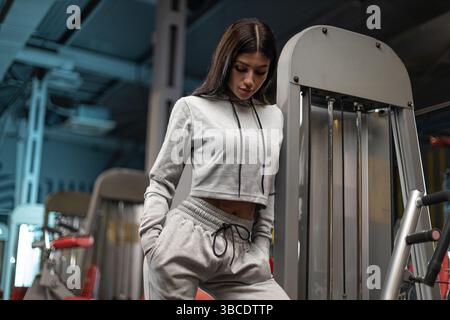 Junge Frau in grauer Sportkleidung steht selbstbewusst in einem modernen Fitnessstudio. Sie scheint konzentriert und gut vorbereitet für ihre Trainingsroutine, umgeben von Trainingsgeräten. Stockfoto