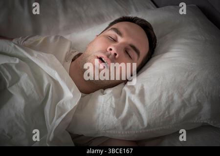 Blick Auf Den Müden Jungen Mann, Der Schnarcht, Während Er Tief Im Bett Schläft. Zum Problem des Schnarchens im Schlaf. Ein junger süßer Kerl schläft nachts auf einem weißen Bett Stockfoto