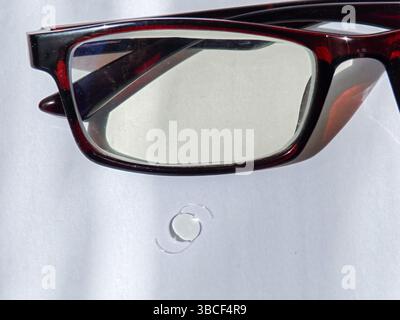 Eine Nahaufnahme einer stilvollen roten Brille auf einer weißen Oberfläche mit einem kleinen runden Glasfragment in der Nähe. Stockfoto