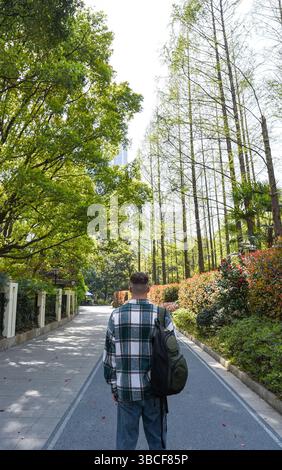 Junger Tourist im People’s Park, Shanghai, von hinten gesehen. Rucksack und kariertes Hemd tragen und einen ruhigen Frühlingstag zwischen Bäumen und Blumen genießen Stockfoto
