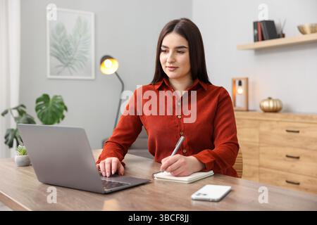 Schöne Frau mit Planer, der Notizen macht und einen Laptop am Holztisch im Haus benutzt Stockfoto
