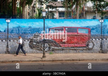 Ein Indianer geht an einem Wandbild vorbei, das einen alten Oldtimer darstellt; in Colaba Area, Mumbai, Indien Stockfoto