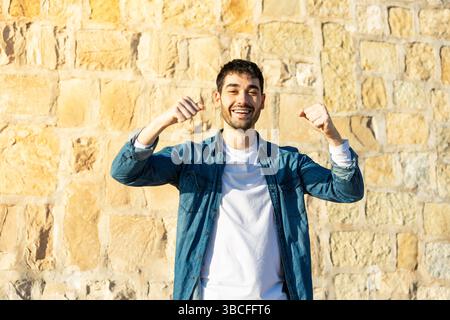 Lächelnder junger Mann in lässigem Jeansshirt, der beide Fäuste zum Feiern hebt, während er draußen vor einem sonnendurchfluteten Steinmauerhintergrund steht. Lifestyle. Stockfoto
