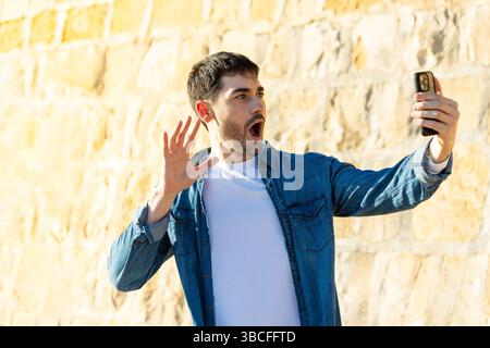 Schockierter junger Mann mit Denim-Hemd und weißem T-Shirt, der das Smartphone auf Armlänge hält, während Videochat im Freien mit breiten Augen und offenem Mund reagiert. Stockfoto