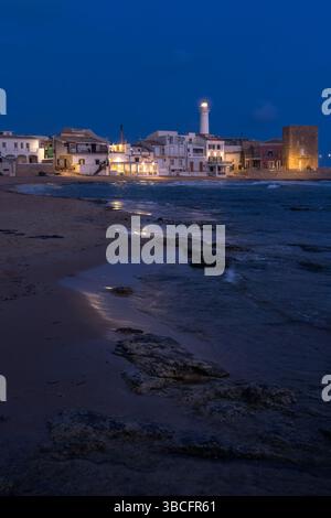 Sanfte Wellen umrunden das Ufer, während warme Lichter auf die historischen Gebäude von Punta Secca leuchten und einen ruhigen Abend in Sizilien, Italien, schaffen Stockfoto
