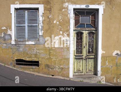 Die Vorderseite eines alten verlassenen gelben Hauses mit blau verschlossenen Fenstern und verschlossener grüner Holztür mit abblätternder Farbe auf einer schrägen Straße Stockfoto