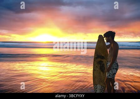 Ein Surfer steht mit Surfbrett in der Hand am Strand und beobachtet den Sonnenaufgang über dem Meer an einem bewölkten Tag, der atemberaubende Farben in den Wolken erzeugt. Stockfoto