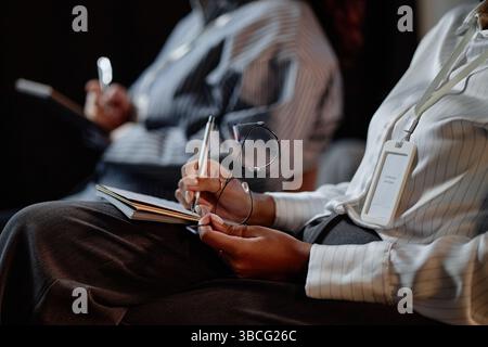 Nahaufnahme einer schwarzen Konferenzteilnehmerin, die eine Brille in der Hand hält, während sie Notizen über die Präsentationen in Notizbuch während des Geschäftsbetriebs hält Stockfoto
