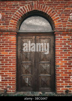 Eine alte Holzdoppeltür in einer roten Ziegelwand mit einem gewölbten Milchfenster darüber, die rustikale europäische Architekturdetails zeigt. Stockfoto