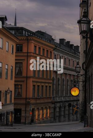 Blick in die Dämmerung auf eine ruhige Straße in Stockholms Altstadt (Gamla Stan) mit historischen Gebäuden, einem leuchtenden Apothekenschild und schwedischer Flagge, Schweden. Stockfoto