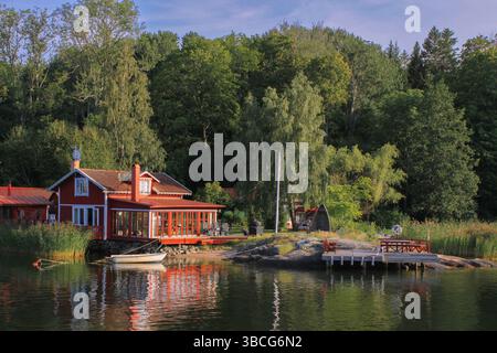 Traditionelles rotes schwedisches Sommerhaus mit Hafenbecken am See und Ruderboot umgeben von üppigen grünen Wäldern im Stockholmer Archipel. Stockfoto