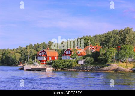 Traditionelle rote Holzhäuser mit Hafenstegen im Stockholmer Archipel, Schweden, umgeben von üppigen grünen Wäldern unter einem Sommerhimmel. Stockfoto
