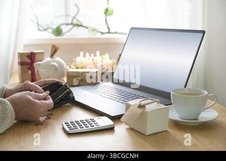 Teure Weihnachtseinkäufe im Internet, Hände einer älteren Frau zählen Geld auf einem Tisch mit Laptop, Geschenke, Kerzen, Schweinebank und Taschenrechner, Kopie sp Stockfoto