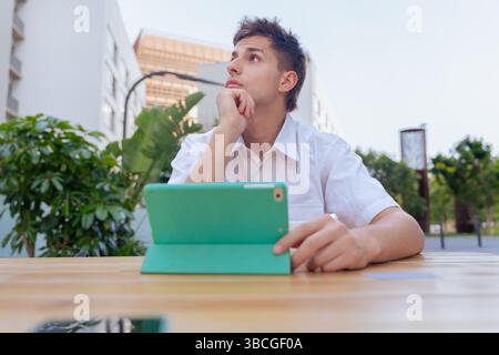 Nachdenklicher junger Mann im Freien mit Tablet und Kaffee in urbaner Umgebung Stockfoto