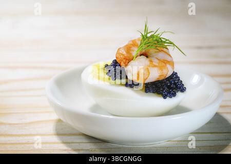 Halbierte gekochte Eier mit schwarzem Kaviar, Garnelen und Dill garnieren auf einem kleinen weißen Teller und einem Holztisch, Fingerfood Snack für Feiertage wie Ostern, CH Stockfoto