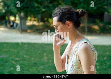 Frau, die auf einem Handy in einem sonnigen Park mit grünem Gras und Bäumen spricht. Brüssel, Belgien Stockfoto