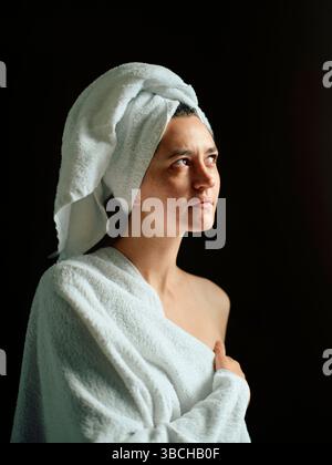Frau in ein Handtuch gewickelt mit einem Handtuch auf dem Kopf vor dunklem Hintergrund. Stockfoto