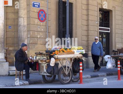 NIKOSIA, ZYPERN, 17. März 2018: Ein Mann, der Obst von einem Stand auf einem Wagen verkauft, sitzt auf dem Bürgersteig in Nikosia Zypern an einer Straßenecke, während ein Mann auf dem Weg geht Stockfoto