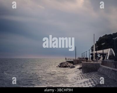 Ein Küstenweg in Grado, Italien, erstreckt sich unter einem stimmungsvollen Himmel ins Meer. Wellen schlagen gegen Steinbarrieren, während ein paar Leute entlang des El spazieren Stockfoto