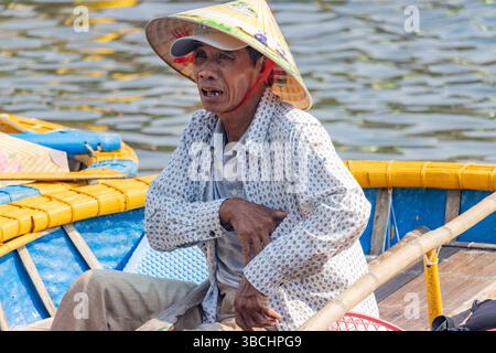HOI AN, VIETNAM, 26. April 2025, Ein Mann mit traditionellem Strohhut und gemustertem Hemd, der auf einem bunten Holzboot sitzt Stockfoto