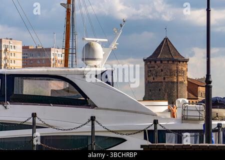 Moderne Yacht, die an einem Yachthafen mit einem historischen Backsteinturm und zeitgenössischen Wohngebäuden im Hintergrund verankert ist. Der Kontrast zwischen traditionellem Stockfoto