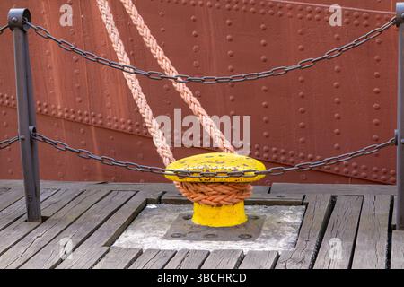Gelber Schiffspoller mit einem dicken Seil, das an den Kai gebunden ist, an den genieteten Rumpf eines Schiffes gesetzt. Hölzerne Dockdielen und eine Sicherheitskette sorgen für eine Instruktion Stockfoto