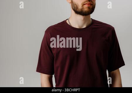 Lässiges Männermodell mit einem burgunderroten T-Shirt vor einem schlichten grauen Hintergrund während eines Fotoshootings im Studio Stockfoto