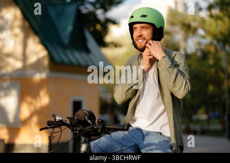 Ein Mann mit grünem Helm passt ihn an, während er im sonnigen Park auf dem Fahrrad sitzt Stockfoto