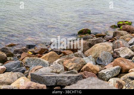Sandstrand mit Felsen und Vegetation im Vordergrund, sanfte Wellen wäschen das Ufer. Natürliche Küstenlandschaft in gedämpften Tönen. Stockfoto