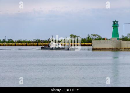 Kleines Boot in der Nähe des Hafeneingangs, mit grünem Leuchtturm und Wellenbrecher im Hintergrund. Eine ruhige maritime Szene mit Hafeninfrastruktur. Stockfoto