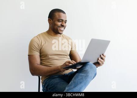 Fröhlicher Schwarzer, Der Laptop Benutzt, Online Arbeitet, Während Er Drinnen Auf Dem Stuhl Sitzt Stockfoto