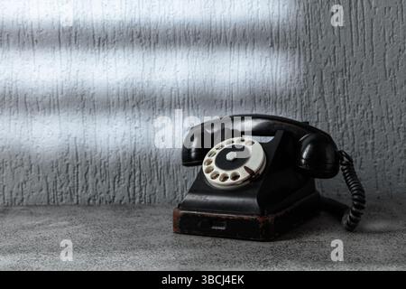Vintage-schwarzes Telefon mit Drehknopf auf grauem Steintisch. Ein Lichtstrahl fällt durch ein Fenster mit Gitter auf die graue Wand. Das Konzept von HO Stockfoto