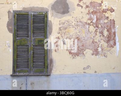 Eine alte, gerissene Hauswand mit geflicktem Zement, lackiert in abblätternden Schichten aus grau-gelber und roter Farbe, mit einem grünen Holzrissfenster mit c Stockfoto